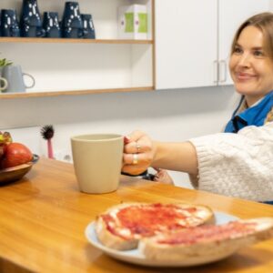 Eine junge weibliche Person, die eine blaue Weste der Bahnhofsmission trägt, reicht eine Tasse über einen Tresen. Sie lächelt ihr Gegenüber an, von dem nur eine Hand zu sehen ist. Auf dem Tresen steht ein Teller mit Obst und ein Teller mit Marmeladenbroten. Die Szene spielt in der Bahnhofsmission.