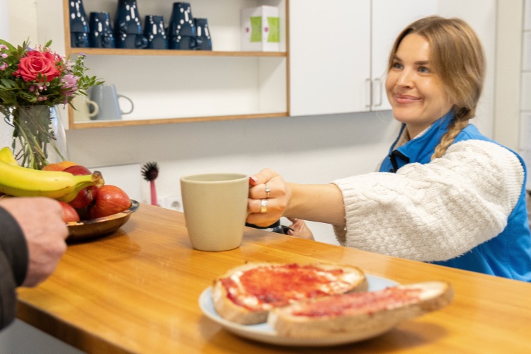 Eine junge weibliche Person, die eine blaue Weste der Bahnhofsmission trägt, reicht eine Tasse über einen Tresen. Sie lächelt ihr Gegenüber an, von dem nur eine Hand zu sehen ist. Auf dem Tresen steht ein Teller mit Obst und ein Teller mit Marmeladenbroten. Die Szene spielt in der Bahnhofsmission.
