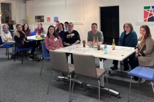 Menschen sitzen in einem Raum an Tischen. Auf den Tischen sind Getränke und Spielkarten zu sehen.