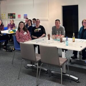 Menschen sitzen in einem Raum an Tischen. Auf den Tischen sind Getränke und Spielkarten zu sehen.