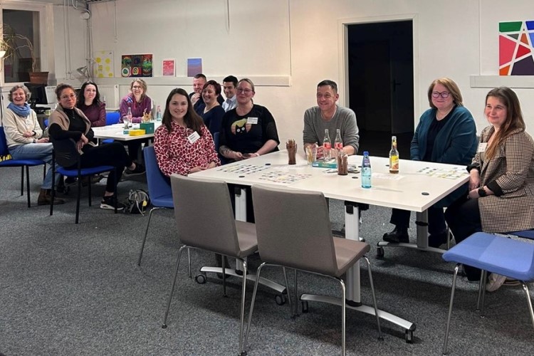 Menschen sitzen in einem Raum an Tischen. Auf den Tischen sind Getränke und Spielkarten zu sehen.